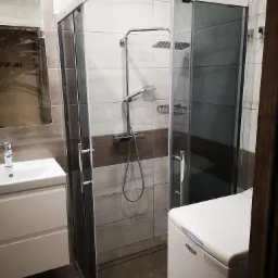 Nowoczesna kabina prysznicowa z przesuwnymi drzwiami, chromowanymi profilami i deszczownicą zamontowana w wyremontowanej łazience z umywalką i pralką.