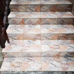 Schody zewnętrzne wyłożone płytkami o nieregularnym wzorze w odcieniach beżu, brązu i szarości, z drewnianą barierką po lewej stronie i kamienną ścianą po prawej.