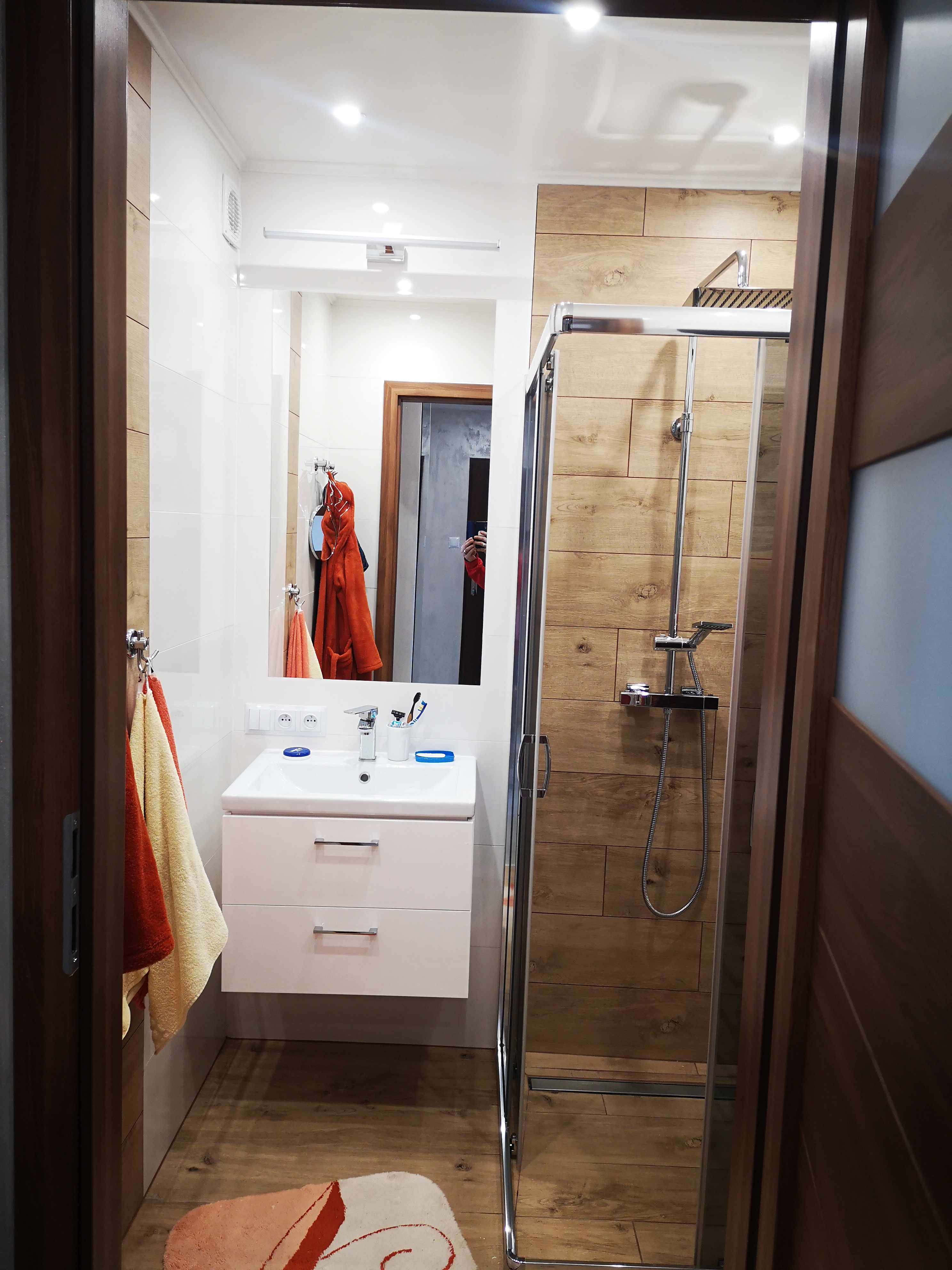 Nowoczesna łazienka z kabiną prysznicową typu walk-in, wykończona płytkami imitującymi drewno na ścianie prysznica i podłodze, biała umywalka z szafką, lustro z oświetleniem i ręczniki w odcieniach...