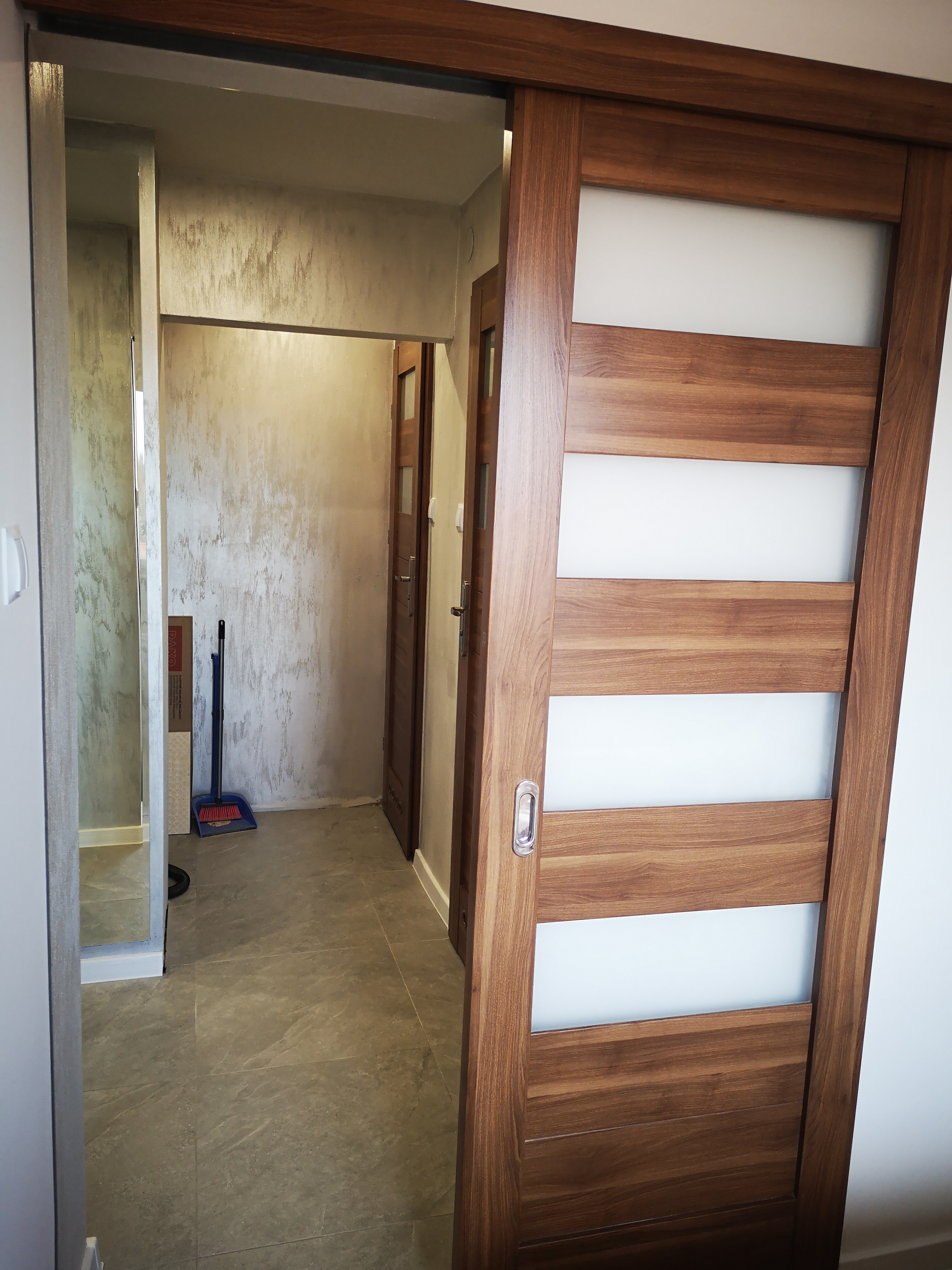 Wnętrze mieszkania w trakcie wykańczania, z widocznymi przesuwnymi drzwiami z drewnianą ramą i matowym szkłem, szarą podłogą z płytek imitujących kamień oraz ścianą z efektem dekoracyjnym; w tle...