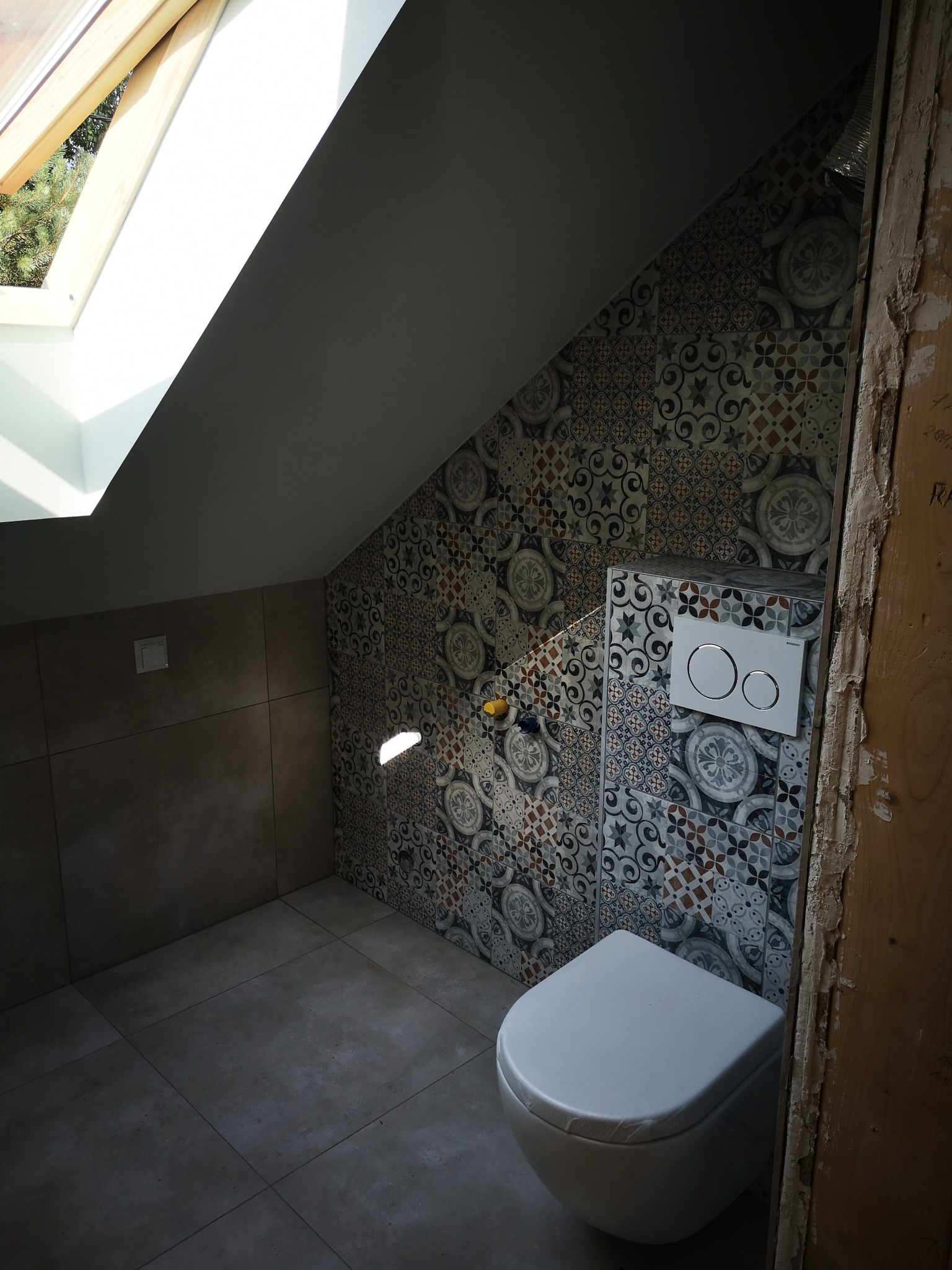 Wykończona łazienka na poddaszu ze skośnym sufitem, okno dachowe wpuszcza naturalne światło, widoczna toaleta wisząca z ozdobnymi płytkami na ścianie i brązowe płytki na podłodze i ścianie po lewej...