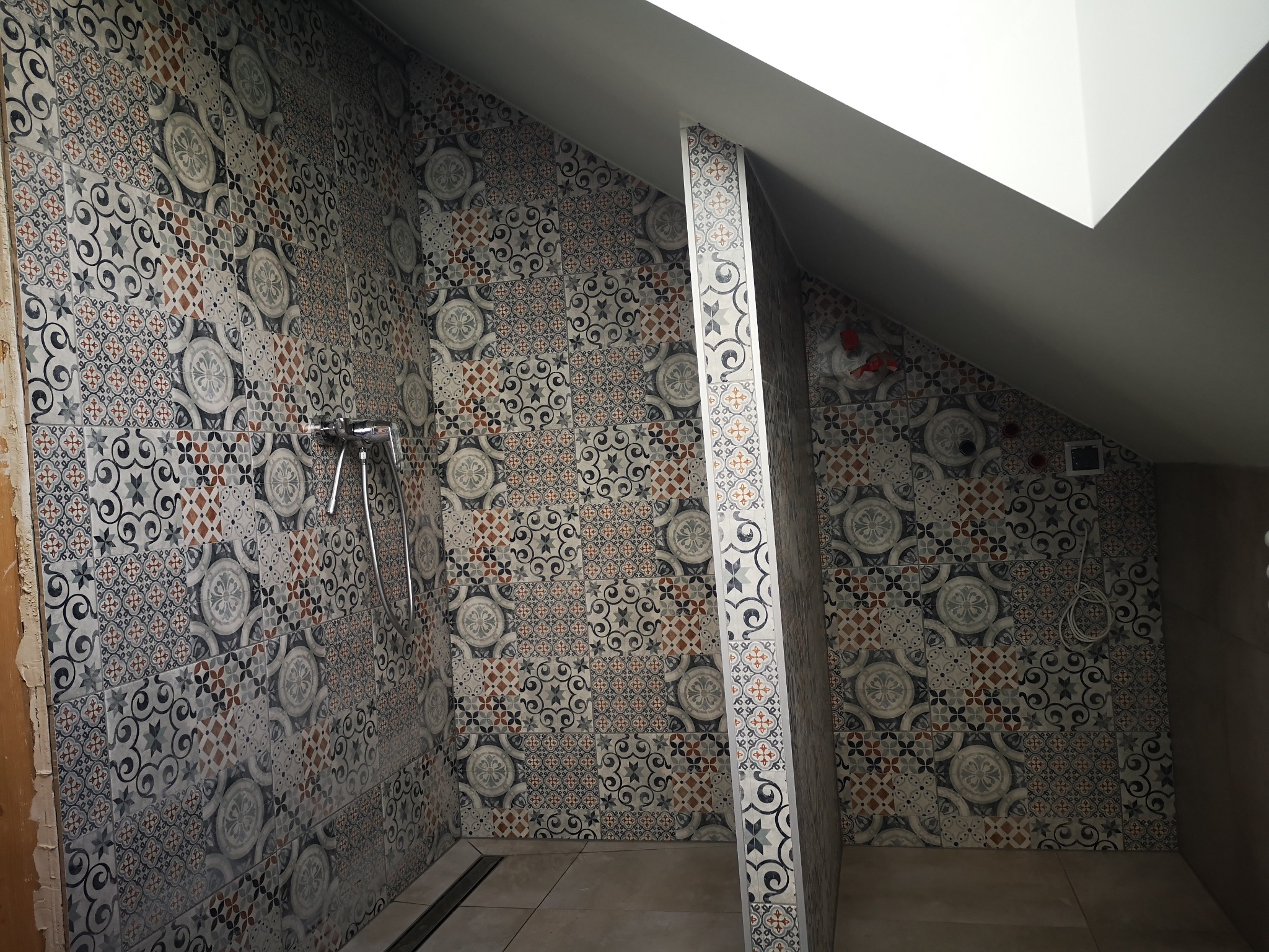 Wykończona kabina prysznicowa na poddaszu z wzorzystymi, kolorowymi płytkami ceramicznymi na ścianach i kolumnie, widoczna bateria prysznicowa i odpływ liniowy w podłodze.