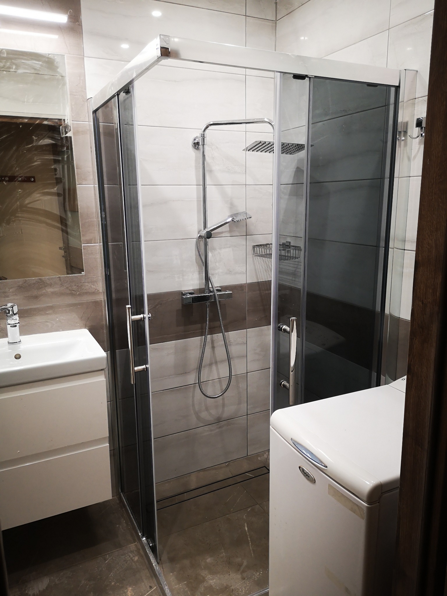 Nowoczesna kabina prysznicowa z przesuwnymi drzwiami, chromowanymi profilami i deszczownicą zamontowana w wyremontowanej łazience z umywalką i pralką.