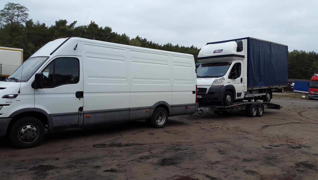 Biały bus holuje na lawecie inny biały samochód dostawczy z niebieską plandeką na tle drzew.