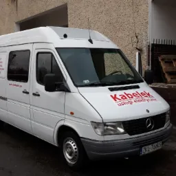 Biały samochód dostawczy firmy świadczącej usługi elektryczne, z czerwonym logo 'Kabelek' na masce i boku, zaparkowany przed budynkiem w pochmurny dzień.