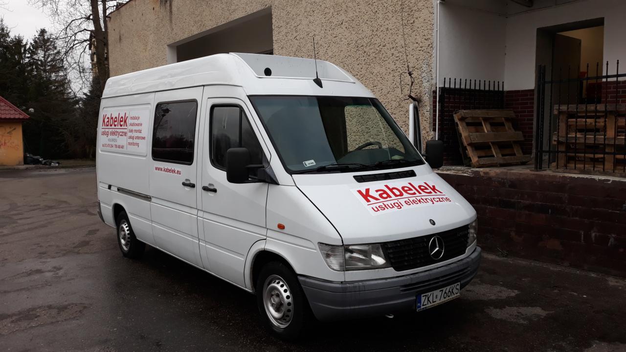 Biały samochód dostawczy firmy świadczącej usługi elektryczne, z czerwonym logo 'Kabelek' na masce i boku, zaparkowany przed budynkiem w pochmurny dzień.