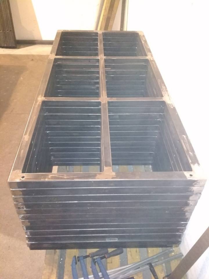 Stos metalowych ram okiennych z widocznymi spawami, ułożonych na palecie, z narzędziami ślusarskimi leżącymi obok.