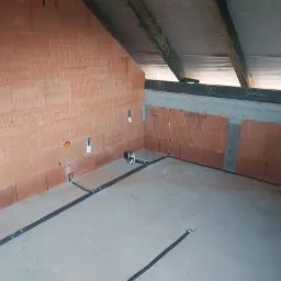 Wnętrze poddasza w trakcie prac wykończeniowych z widocznymi, czarnymi przewodami elektrycznymi ułożonymi wzdłuż ścian z cegły dziurawki.