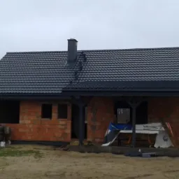 Nowy, grafitowy dach z blachodachówki na surowym budynku z czerwonej cegły, z czarnym kominem i drewnianą konstrukcją werandy, widoczne rusztowanie w tle.