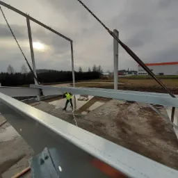 Pracownik w kamizelce odblaskowej montuje stalową konstrukcję hali, widoczne elementy mocujące i łańcuchy dźwigu, w tle budynek magazynowy i pochmurne niebo.