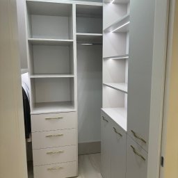 Lepsze Meble - Jasna garderoba z oświetleniem LED, szufladami z złotymi uchwytami i półkami. Widok z perspektywy wejścia do pomieszczenia, ukazujący funkcjonalne wnętrze.