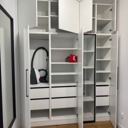 Lepsze Meble - Biała szafa wnękowa z otwartymi drzwiami, widoczne półki, szuflady, odkurzacz i czerwony pojemnik. Minimalistyczny design, drewniana podłoga.