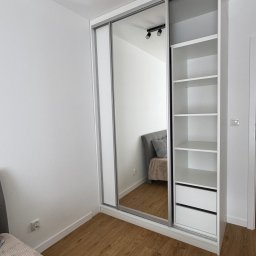 Lepsze Meble - Biała szafa wnękowa z lustrzanymi drzwiami przesuwnymi i otwartymi półkami, widoczna w jasnym wnętrzu sypialni z drewnianą podłogą.