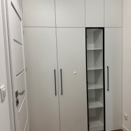 Lepsze Meble - Biała szafa wnękowa z czarnymi, długimi uchwytami i przeszkloną witryną z półkami. Widoczna podłoga z paneli i fragment białej ściany.