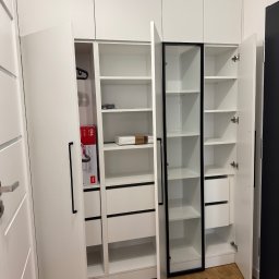 Lepsze Meble - Biała szafa wnękowa z czarnymi akcentami: uchwyty, obramowanie szklanych drzwi. Otwarta, ukazująca półki i szuflady. W środku widoczny odkurzacz w pudełku.