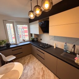 Lepsze Meble - Nowoczesna kuchnia na wymiar z beżowymi frontami, czarnym blatem i marmurową ścianą. Nad blatem wiszą trzy szklane lampy. Widok na blok z okna.