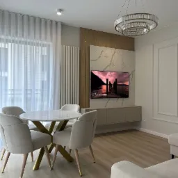 Elegancka aranżacja salonu: okrągły stół z marmurowym blatem i złotą podstawą, krzesła tapicerowane w beżu, ściana TV z marmurowym dekorem i złoty żyrandol.