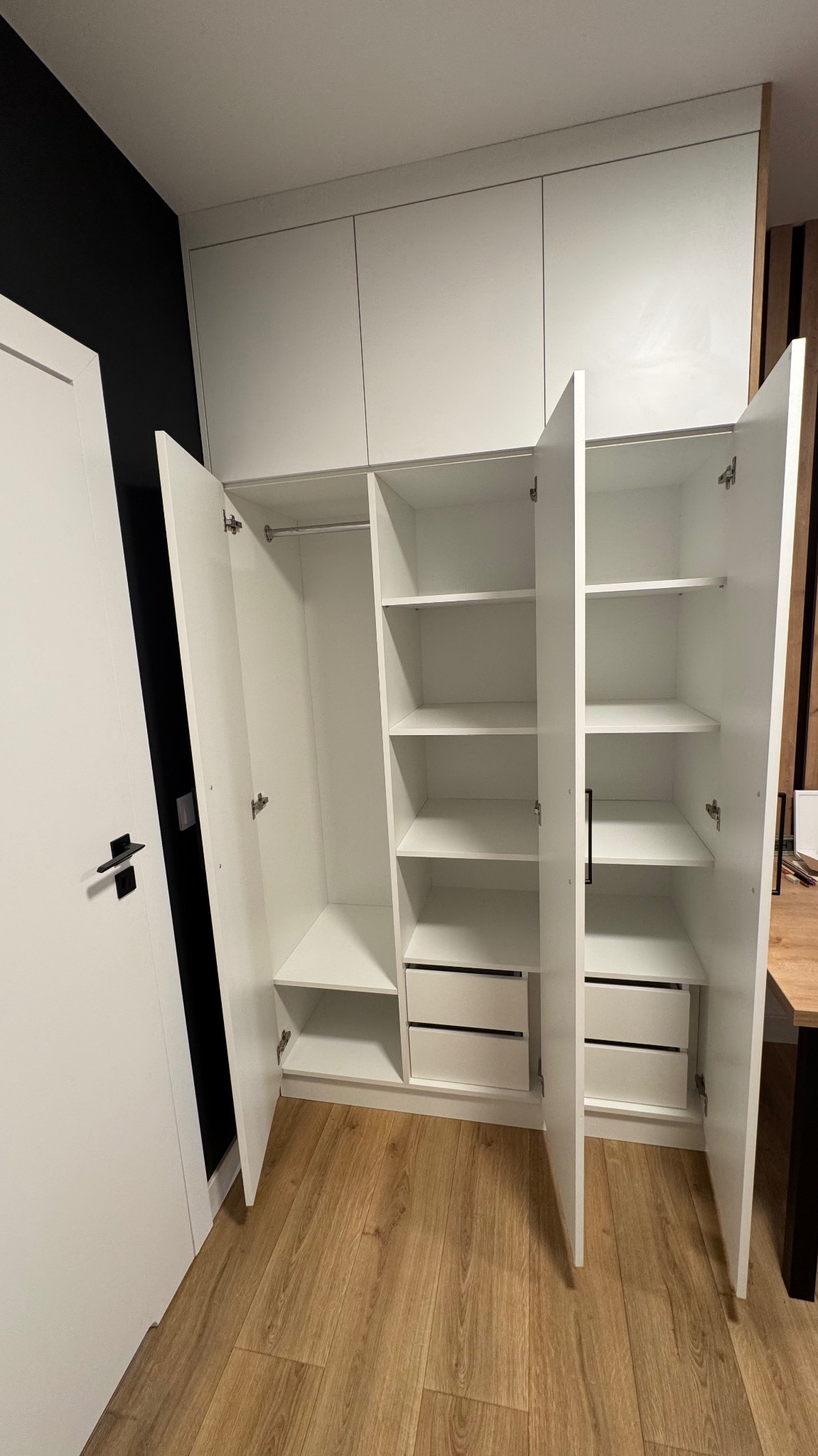 Biała szafa wnękowa z otwartymi drzwiami, widoczne półki, szuflady i drążek na ubrania. Kontrast czarnej ściany i drewnianej podłogi.
