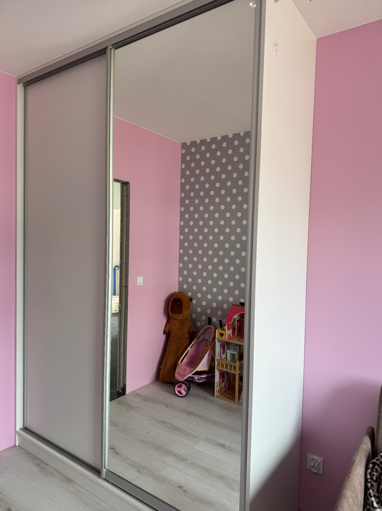 Biała szafa wnękowa z przesuwnymi drzwiami z lustrem, odbijająca pokój dziecięcy z zabawkami i różowo-szarą tapetą w kropki. Estetyczne i funkcjonalne rozwiązanie.