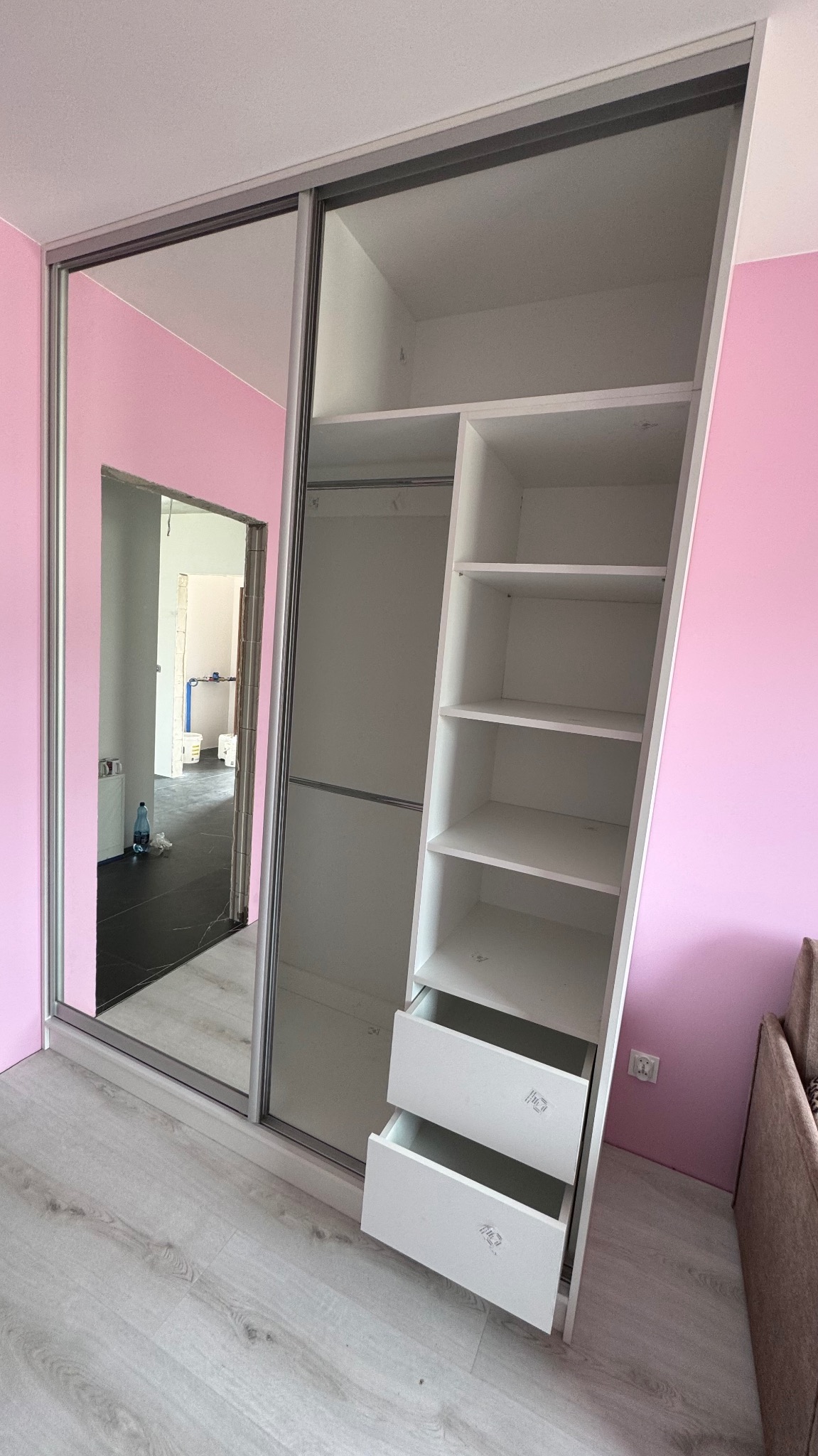 Biała szafa wnękowa z lustrzanymi drzwiami przesuwnymi, z półkami i szufladami, na tle różowej ściany. Widok na wnętrze szafy i pomieszczenie obok.