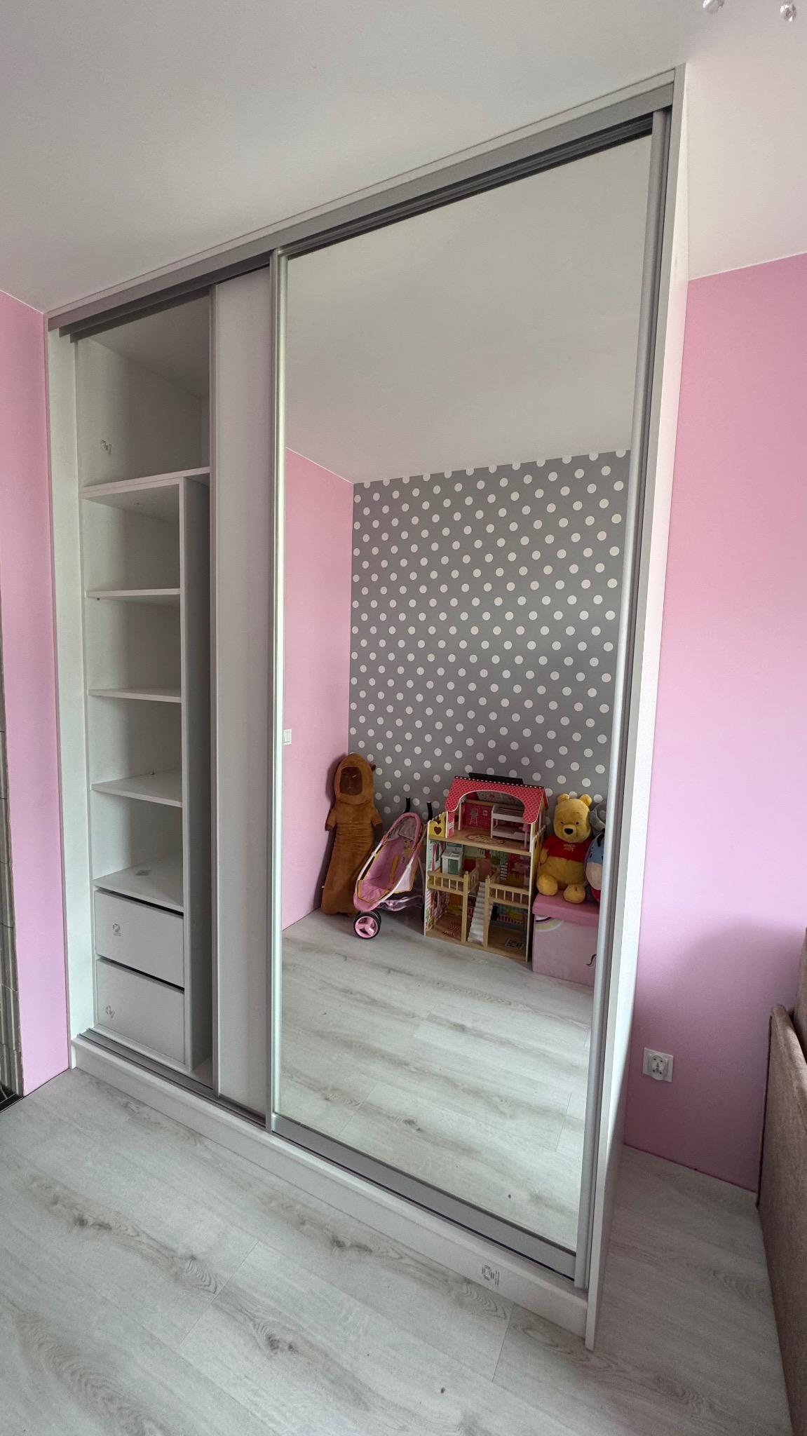 Biała szafa przesuwna z lustrem w pokoju dziecięcym. W lustrze odbicie pokoju z tapetą w kropki, zabawkami i pluszakami. Widoczne półki i szuflady w szafie.