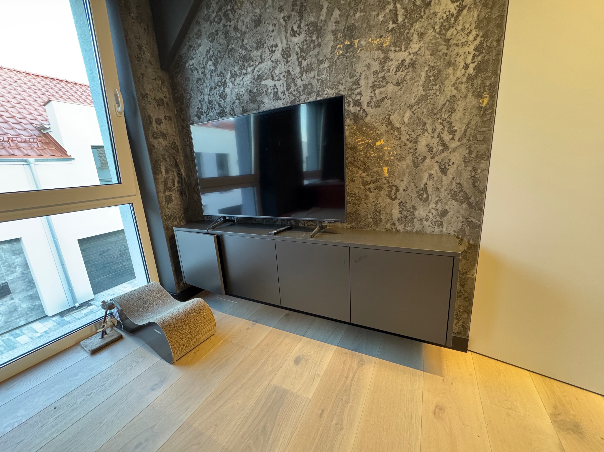 Nowoczesna szafka RTV w odcieniach szarości, z telewizorem na tle ściany o fakturze betonu. Obok okno z widokiem na dom i drapak dla kota.