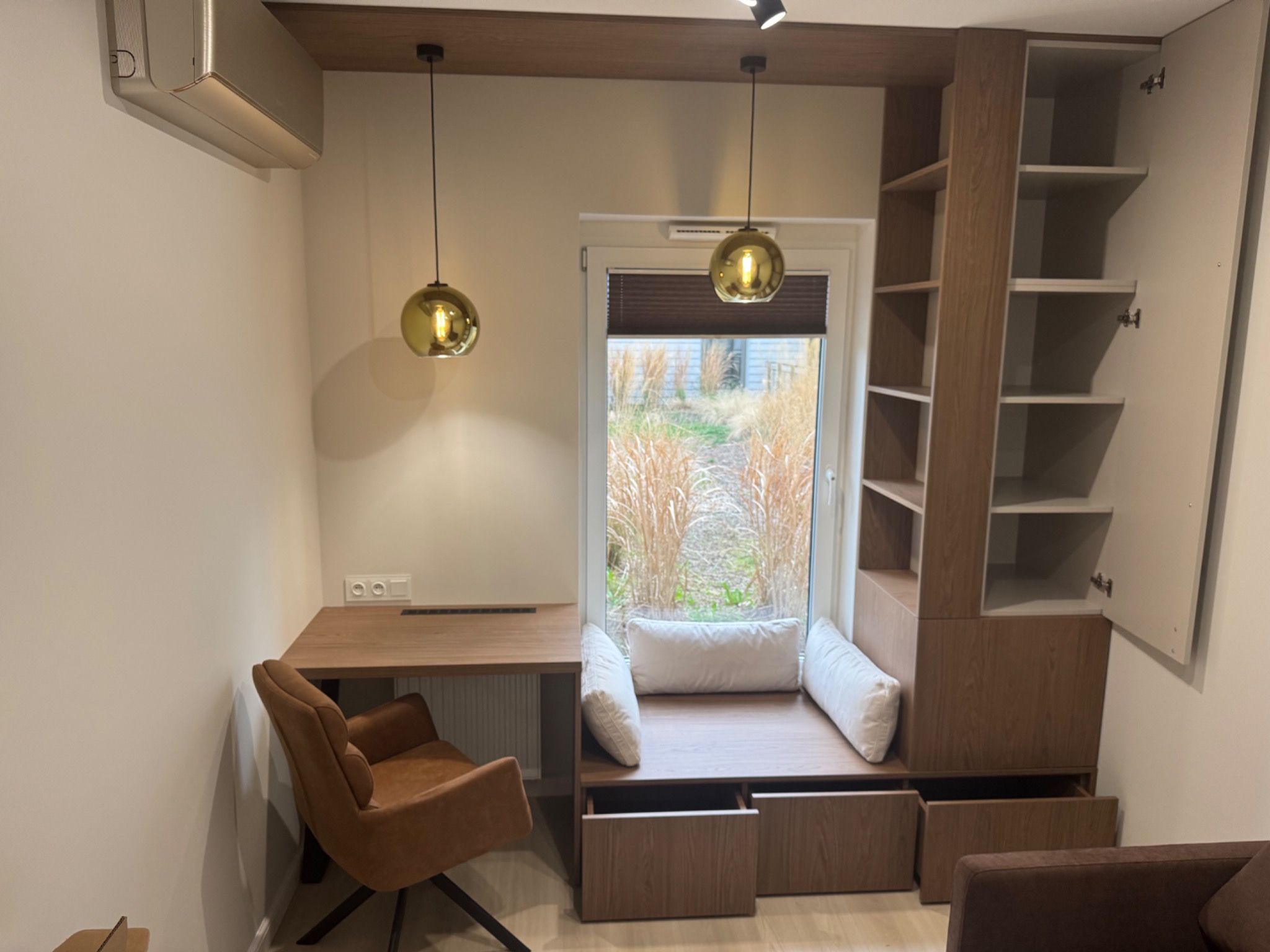 Drewniane biurko z krzesłem, szafa wnękowa i siedzisko przy oknie z widokiem na zewnątrz. Całość w jasnych, naturalnych barwach, dopełniają złote lampy.