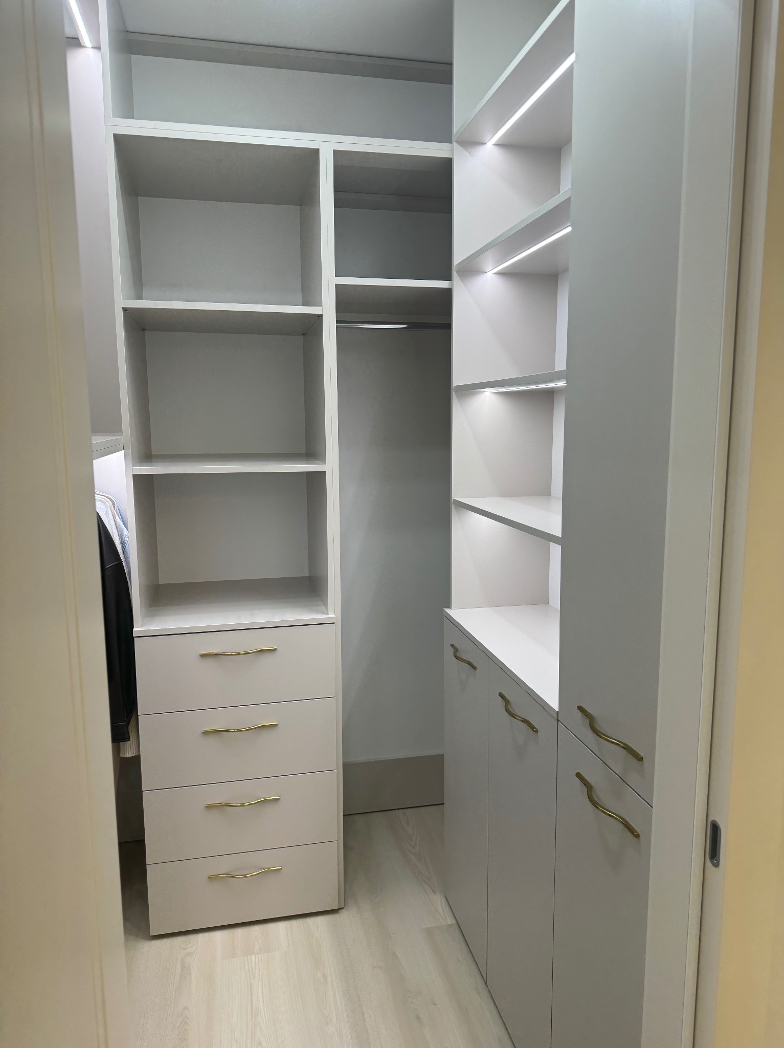 Jasna garderoba z oświetleniem LED, szufladami z złotymi uchwytami i półkami. Widok z perspektywy wejścia do pomieszczenia, ukazujący funkcjonalne wnętrze.