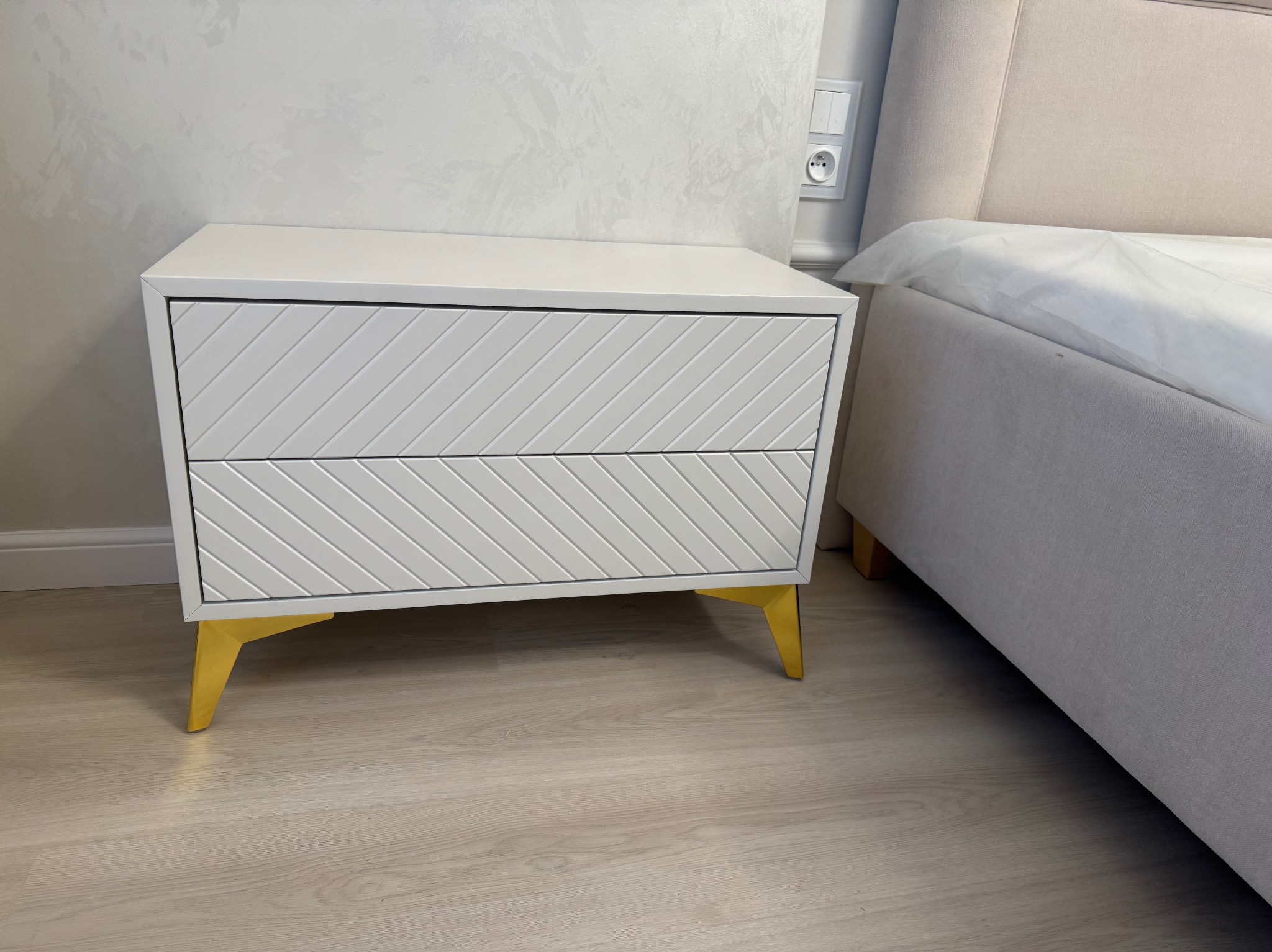 Biała szafka nocna z dwoma szufladami o skośnym wzorze na złotych nóżkach, ustawiona obok łóżka z beżowym zagłówkiem na panelu ściany w tle.
