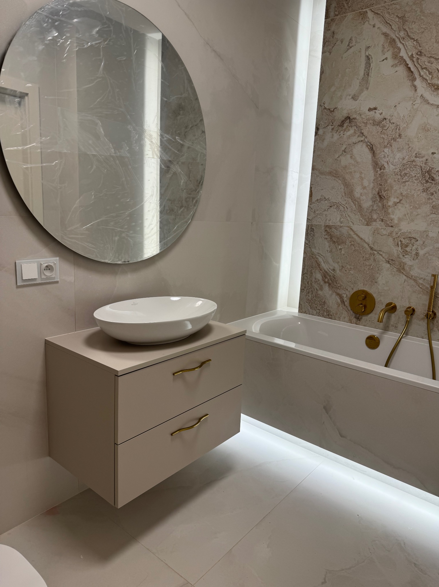 Jasna łazienka z szafką pod umywalkę na wymiar, okrągłe lustro z folią ochronną i wanną z podświetleniem LED. Złota armatura.