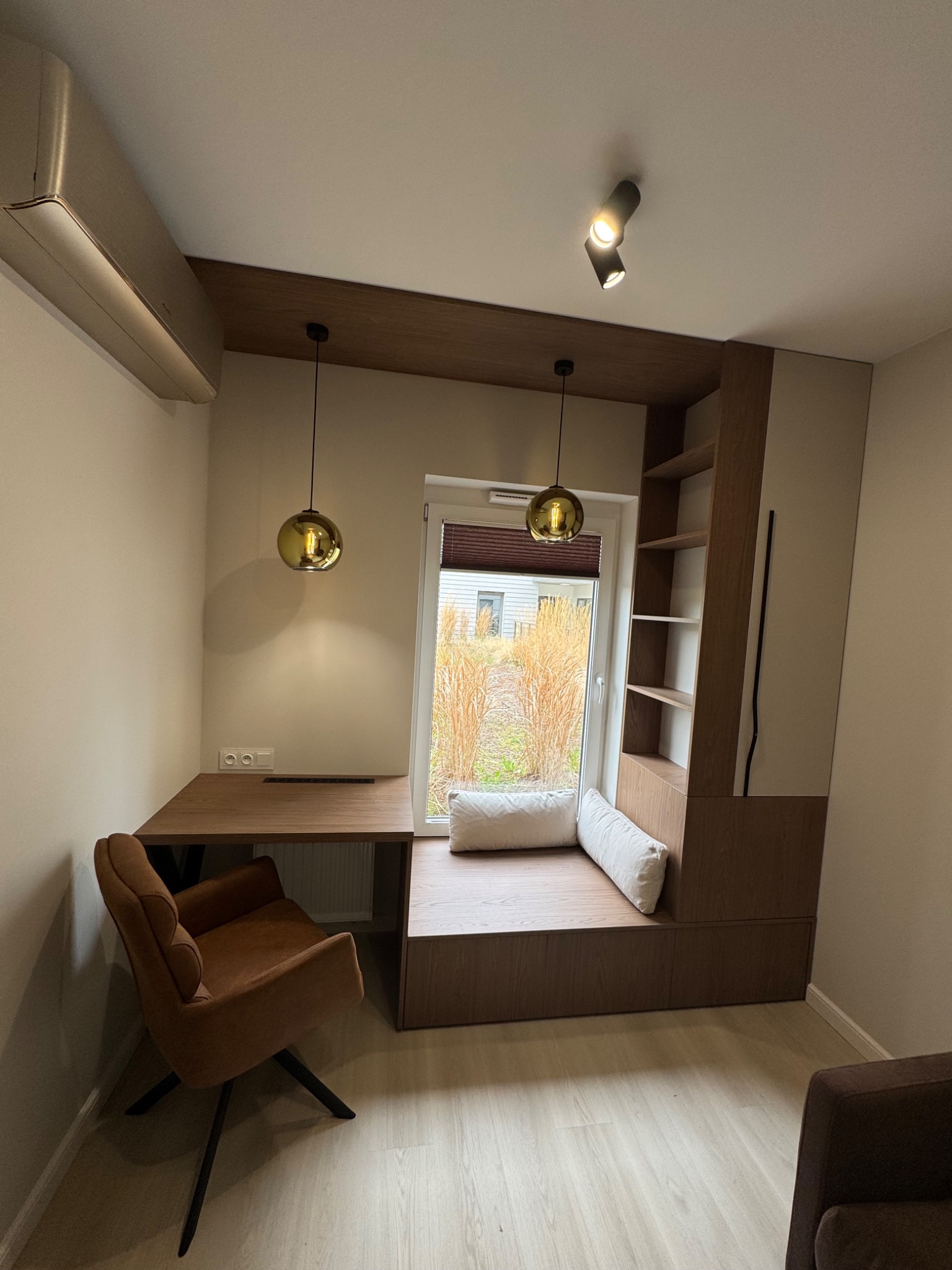 Aranżacja wnętrza z biurkiem, szafką wnękową z półkami i siedziskiem przy oknie. Całość w odcieniach brązu i beżu, dopełniona wiszącymi lampami.