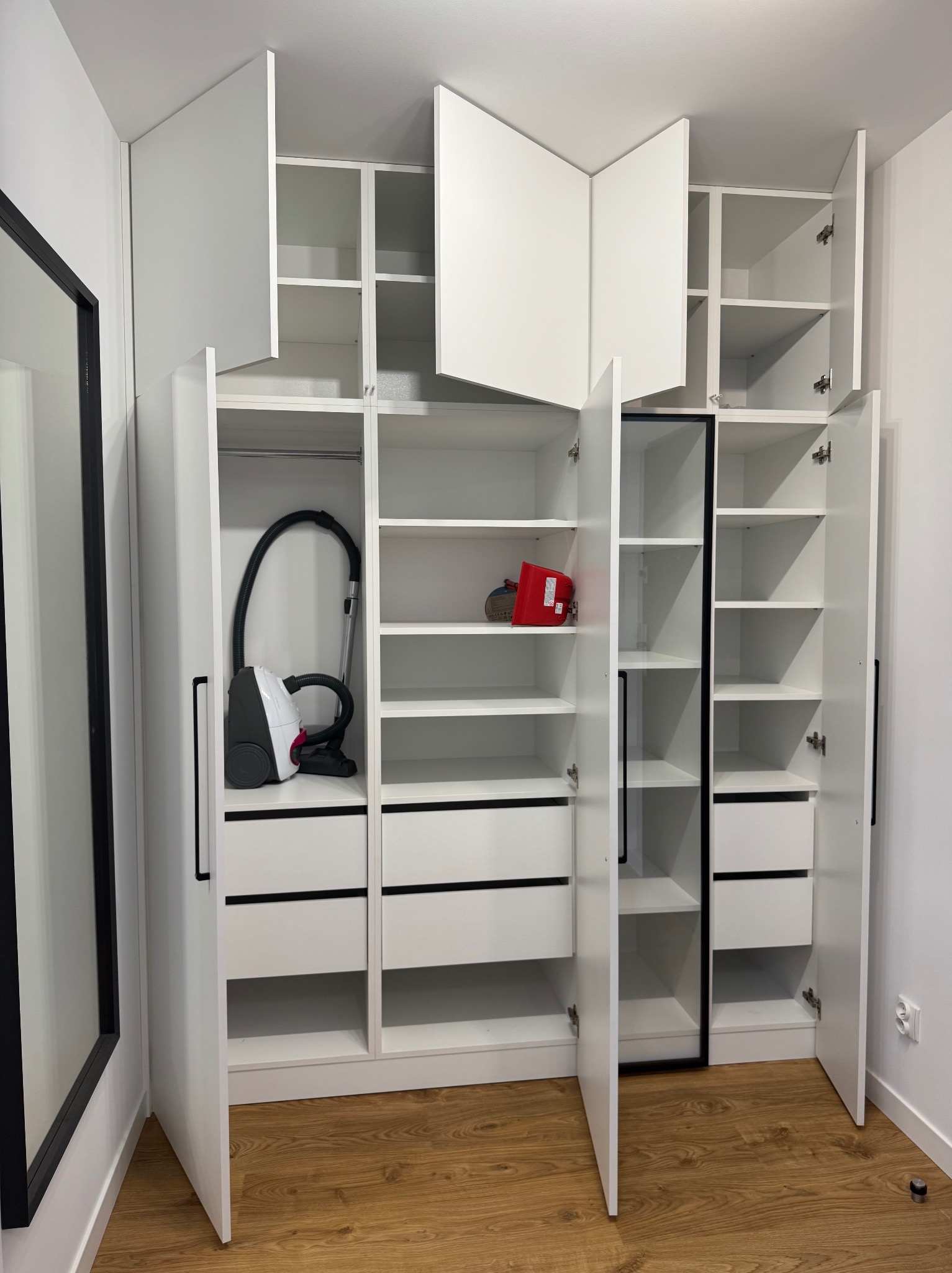 Biała szafa wnękowa z otwartymi drzwiami, widoczne półki, szuflady, odkurzacz i czerwony pojemnik. Minimalistyczny design, drewniana podłoga.