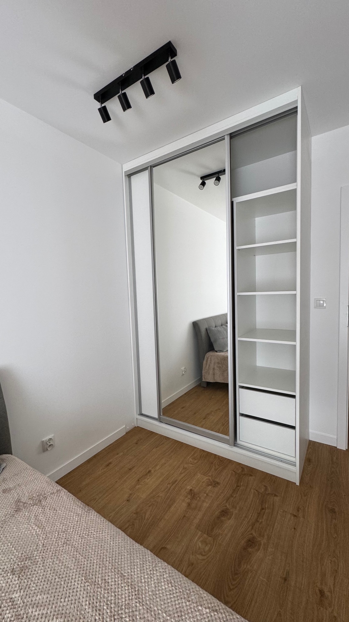 Biała szafa wnękowa z lustrzanymi drzwiami przesuwnymi i otwartymi półkami, widoczna w jasnym wnętrzu sypialni z drewnianą podłogą.