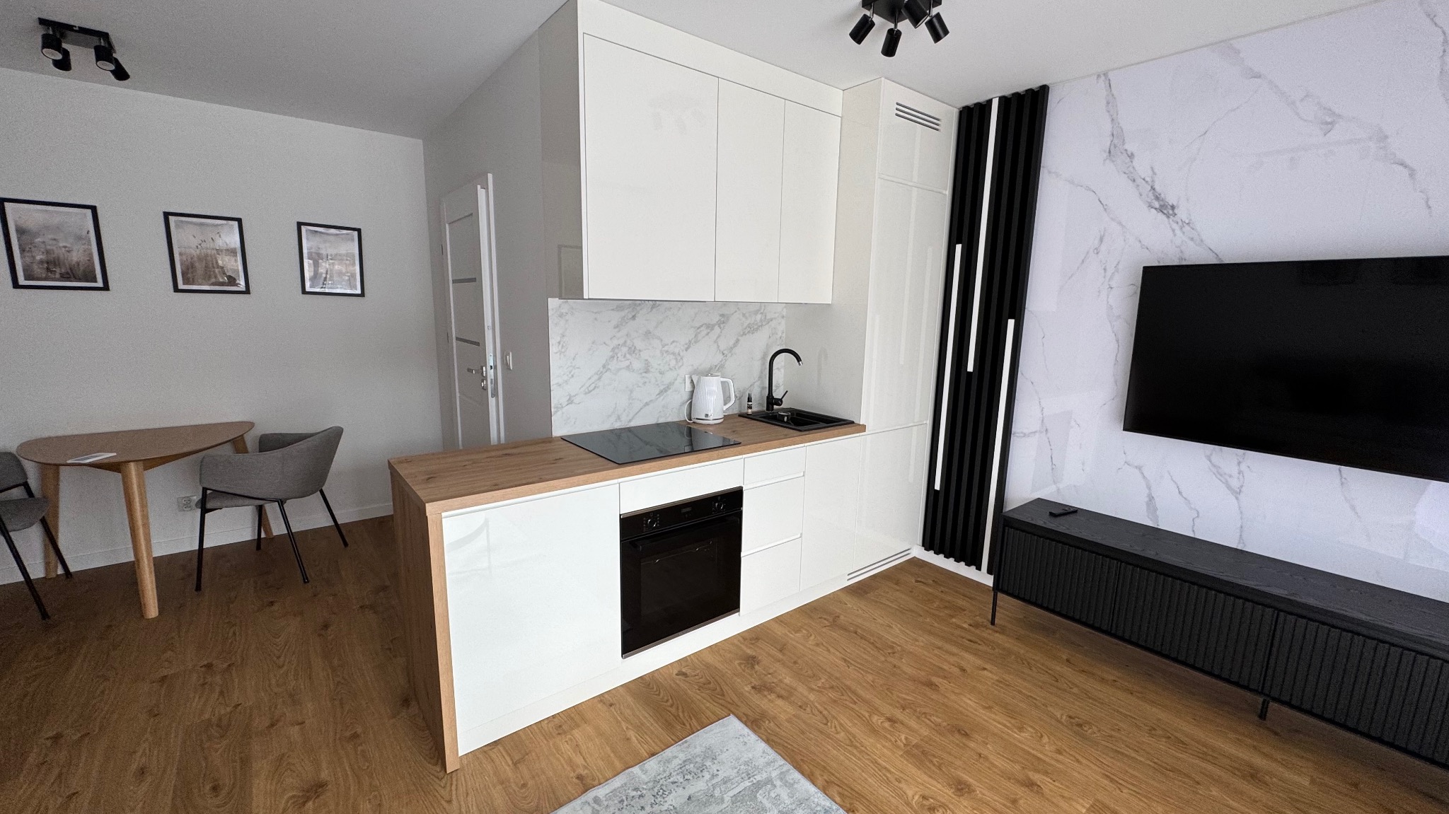 Biała, nowoczesna kuchnia na wymiar z drewnianym blatem i marmurową ścianą. Widoczny stół z krzesłami. Minimalistyczny design, oświetlenie punktowe.