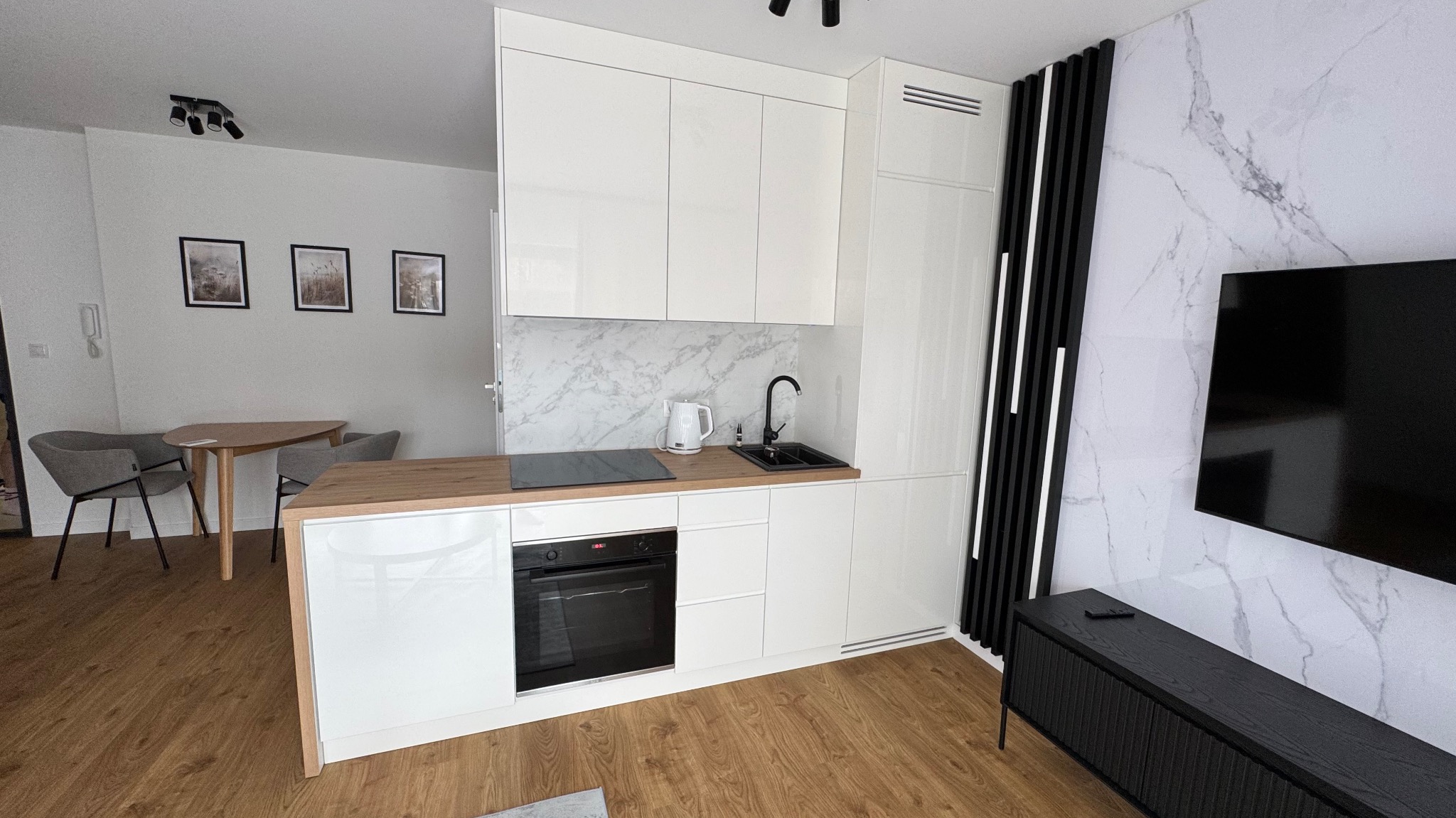 Biała kuchnia na wymiar z drewnianym blatem w nowoczesnym apartamencie. Marmurowe wykończenie ściany i czarne akcenty dekoracyjne.