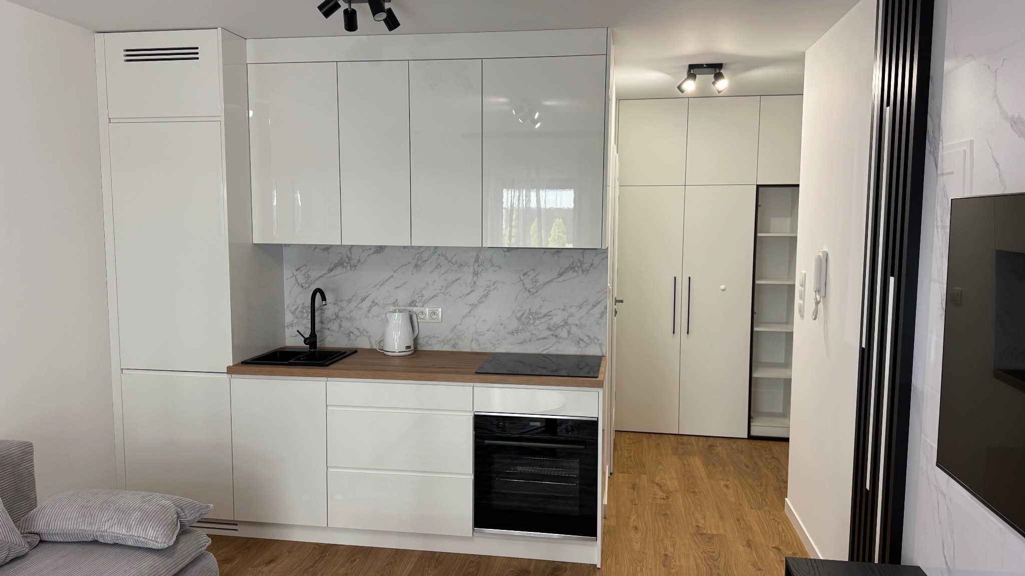 Biała, minimalistyczna kuchnia z marmurowym blatem i czarną armaturą. Widoczna zabudowa szaf wnękowych w przedpokoju, tworząca spójny wystrój wnętrza.