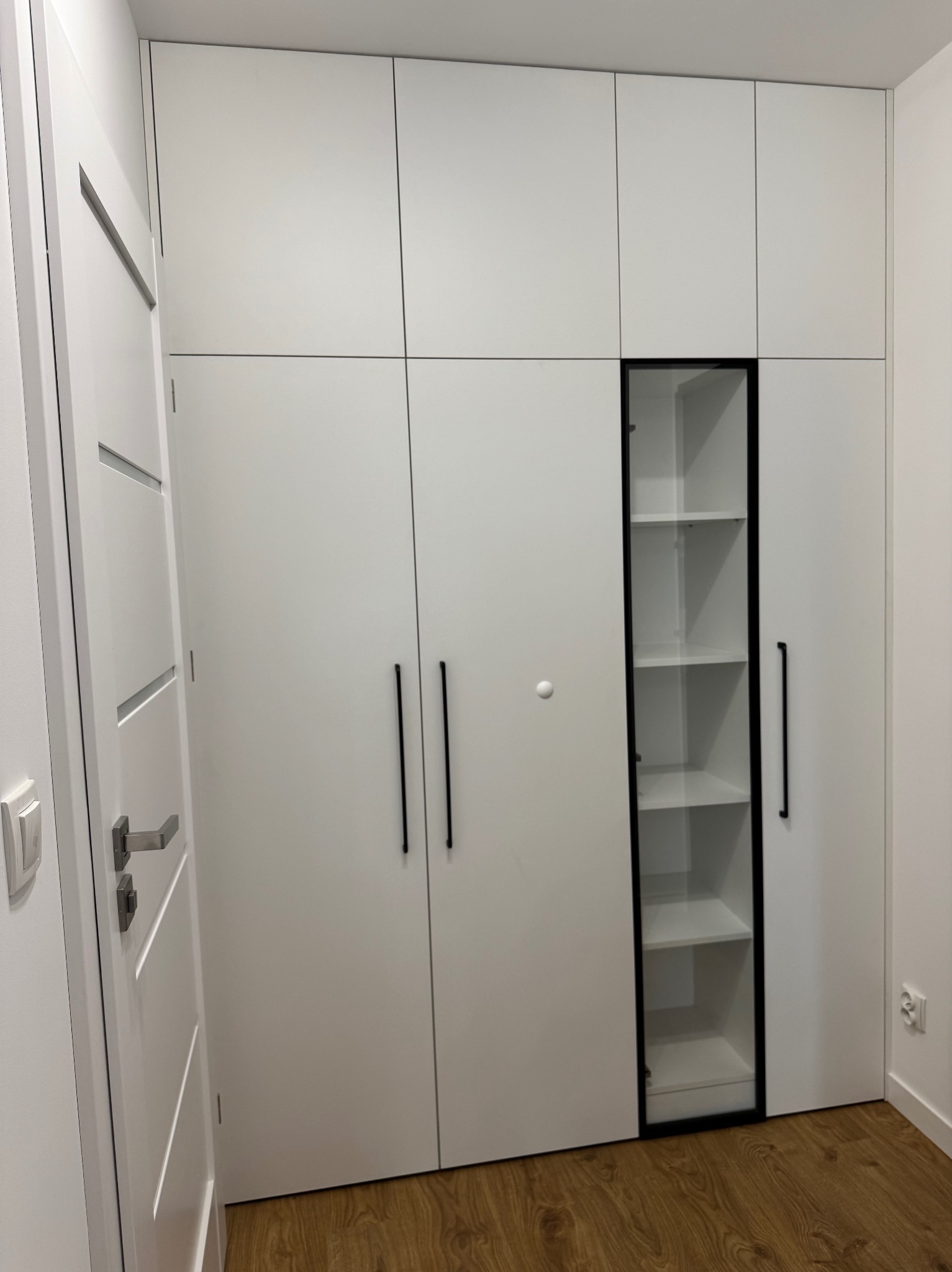 Biała szafa wnękowa z czarnymi, długimi uchwytami i przeszkloną witryną z półkami. Widoczna podłoga z paneli i fragment białej ściany.