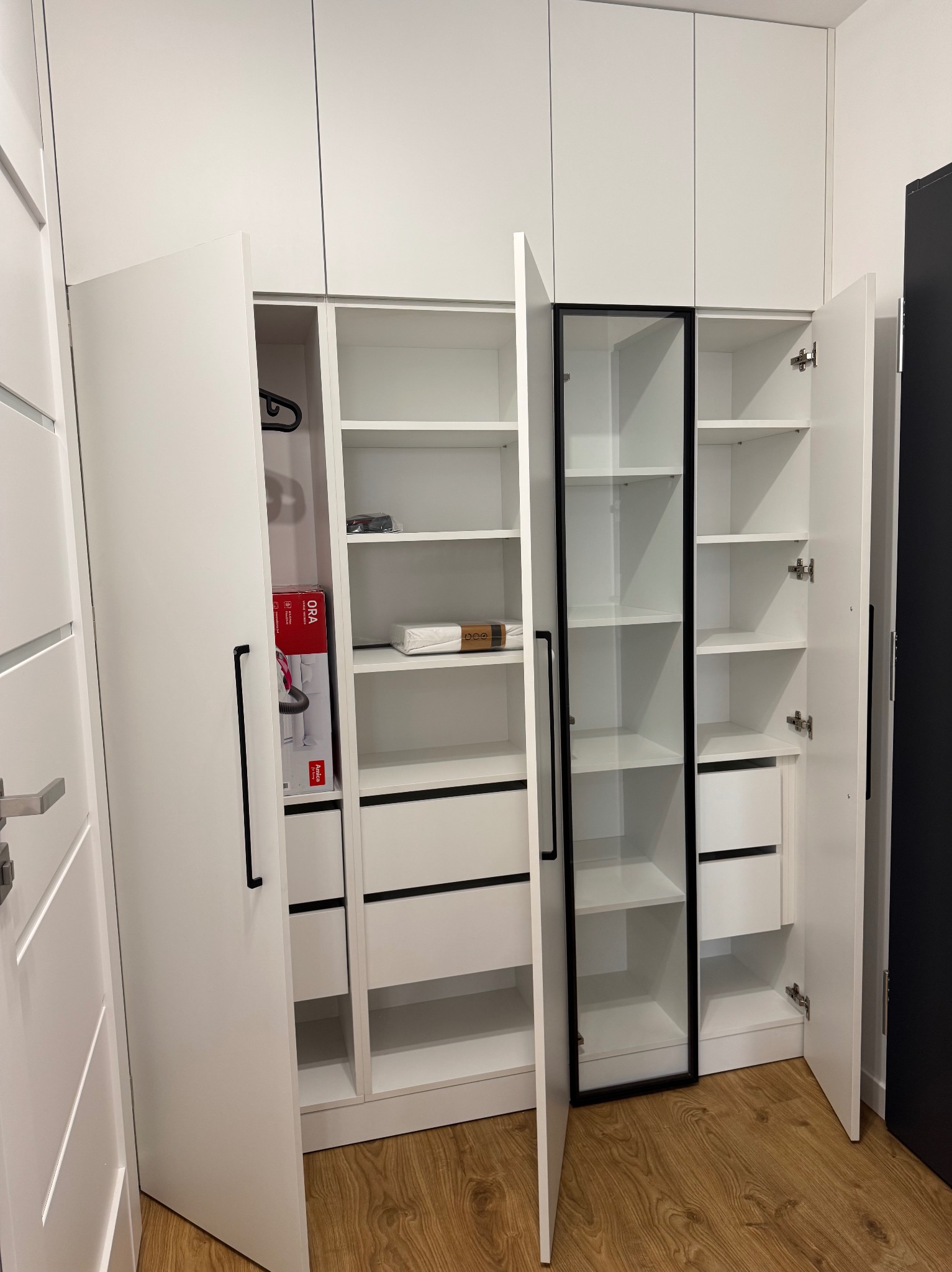 Biała szafa wnękowa z czarnymi akcentami: uchwyty, obramowanie szklanych drzwi. Otwarta, ukazująca półki i szuflady. W środku widoczny odkurzacz w pudełku.