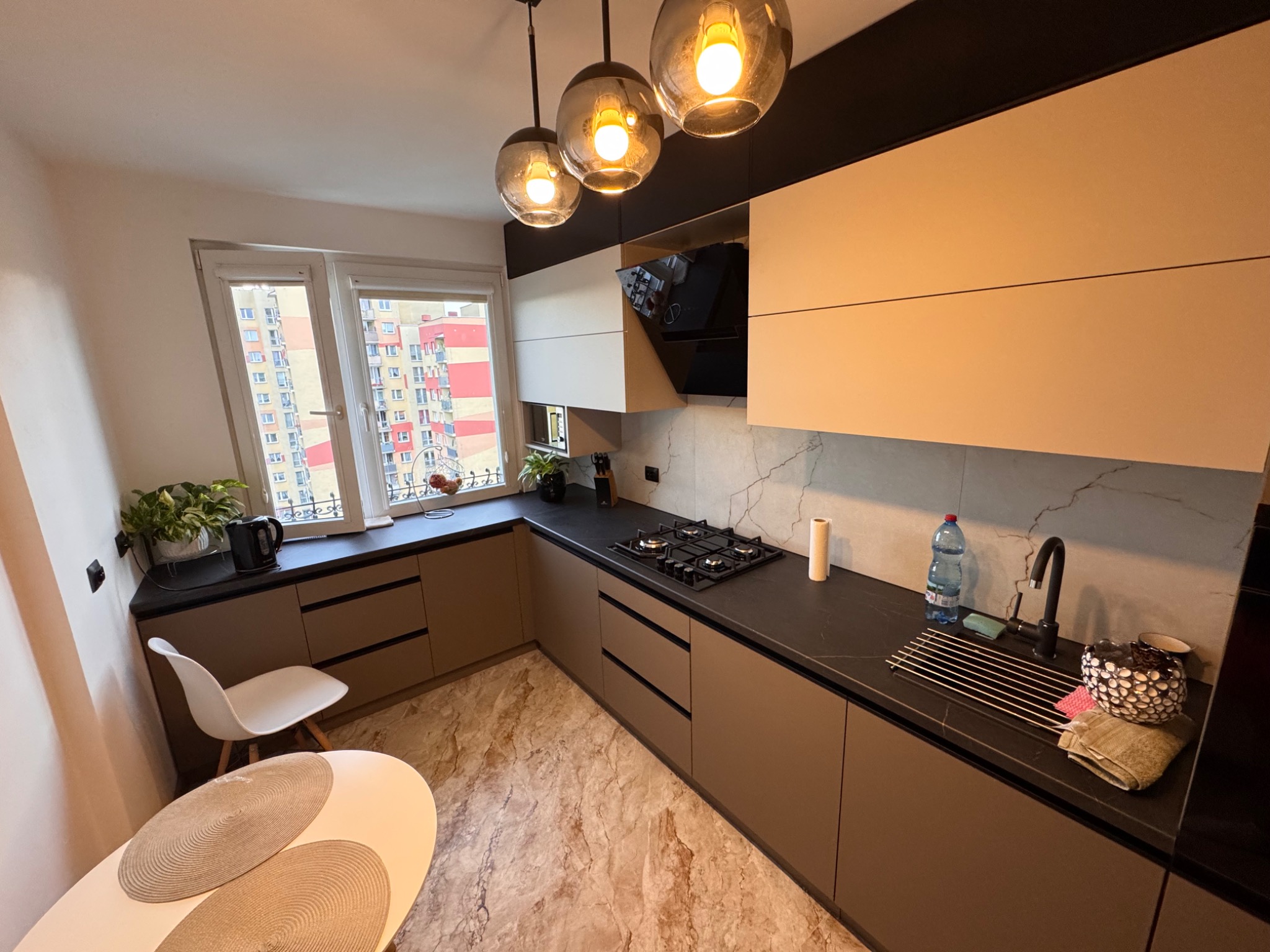 Nowoczesna kuchnia na wymiar z beżowymi frontami, czarnym blatem i marmurową ścianą. Nad blatem wiszą trzy szklane lampy. Widok na blok z okna.
