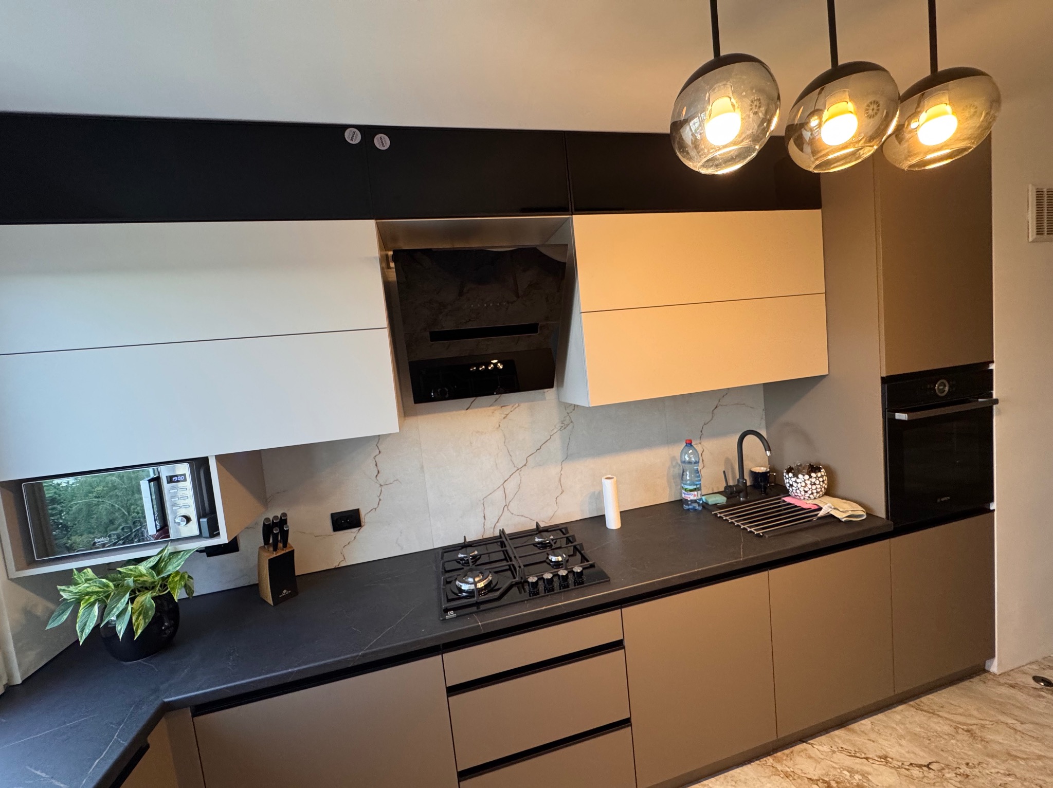 Nowoczesna kuchnia z czarnym blatem, jasnymi szafkami górnymi i brązowymi dolnymi. Nad blatem trzy lampy z okrągłymi kloszami. Na blacie płyta gazowa i akcesoria.