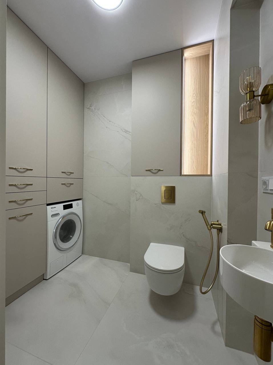 Nowoczesna łazienka z beżowymi szafami, pralką Miele, wiszącą toaletą i złotymi detalami: armaturą, oświetleniem. Minimalistyczny design, płytki imitujące marmur.