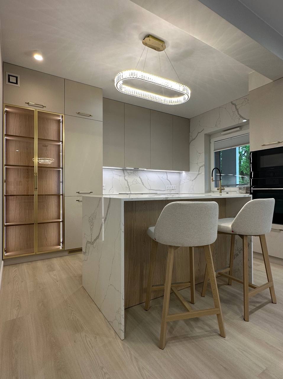 Elegancka kuchnia z wyspą i marmurowym blatem, oświetlona nowoczesną lampą. Dwie szare, drewniane hokery przy wyspie. Szafka z przeszklonymi drzwiami z podświetleniem.