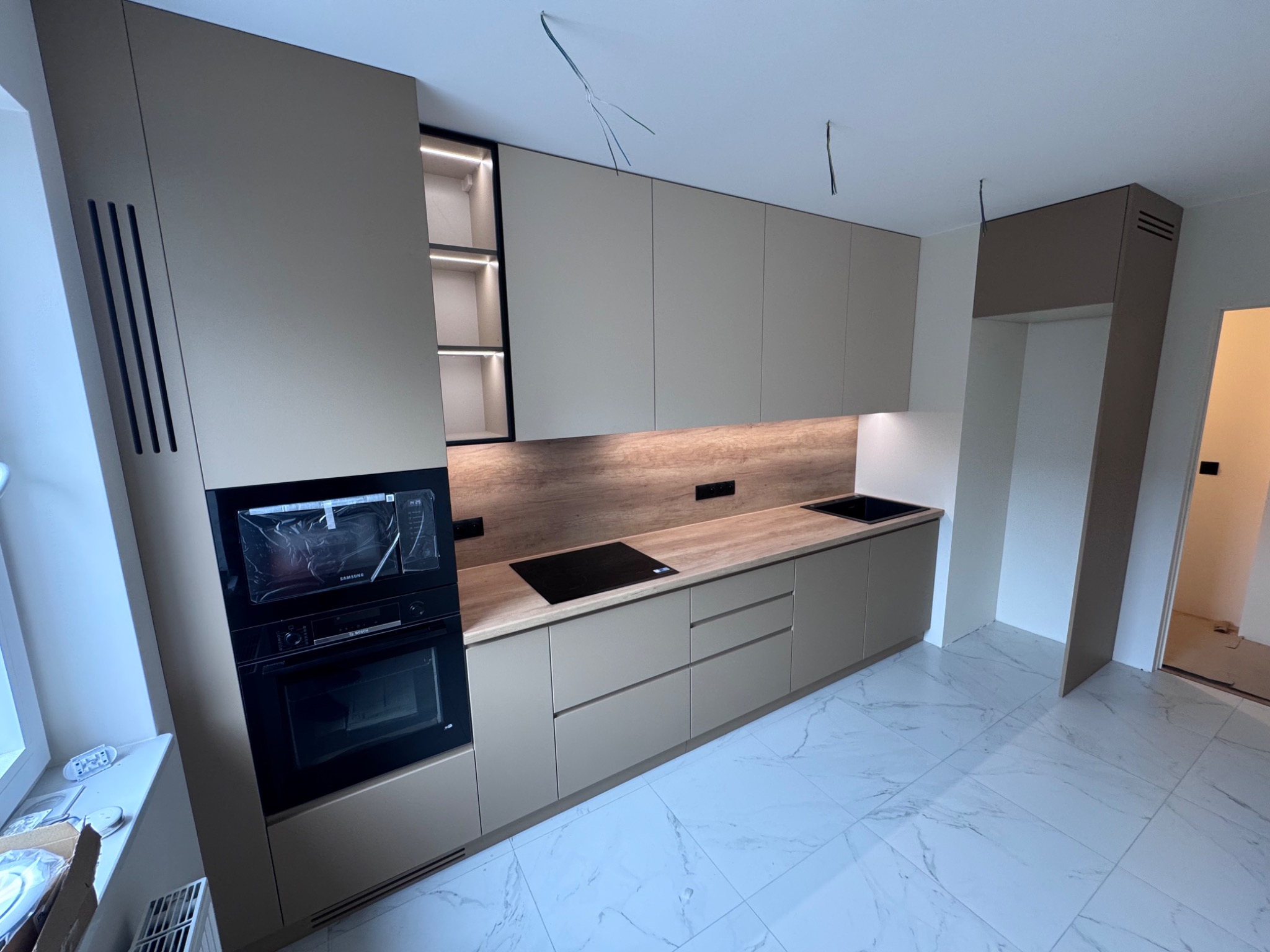 Beżowa kuchnia na wymiar z oświetleniem LED, piekarnikiem, płytą indukcyjną i marmurową podłogą. Uwagę zwracają minimalistyczne fronty i brak uchwytów.