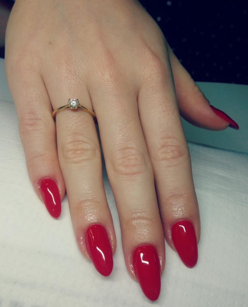 Dłoń z czerwonymi, błyszczącymi paznokciami w kształcie migdała, ozdobiona złotym pierścionkiem z pojedynczym kamieniem, ułożona na białym podłożu.
