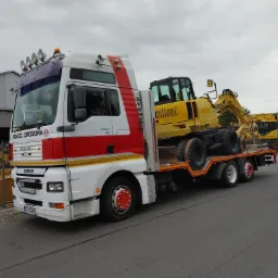 Czerwono-biała ciężarówka MAN z napisem 'Pomoc Drogowa' przewozi żółtą koparkę kołową marki Allrad na niskopodwoziowej naczepie; w tle zaparkowane inne koparki.