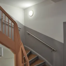 Klatka schodowa w budynku mieszkalnym, widoczne drewniane schody z brązową balustradą, okno z widokiem na zewnątrz oraz pomalowane na szaro ściany z metalową poręczą i lampą sufitową.