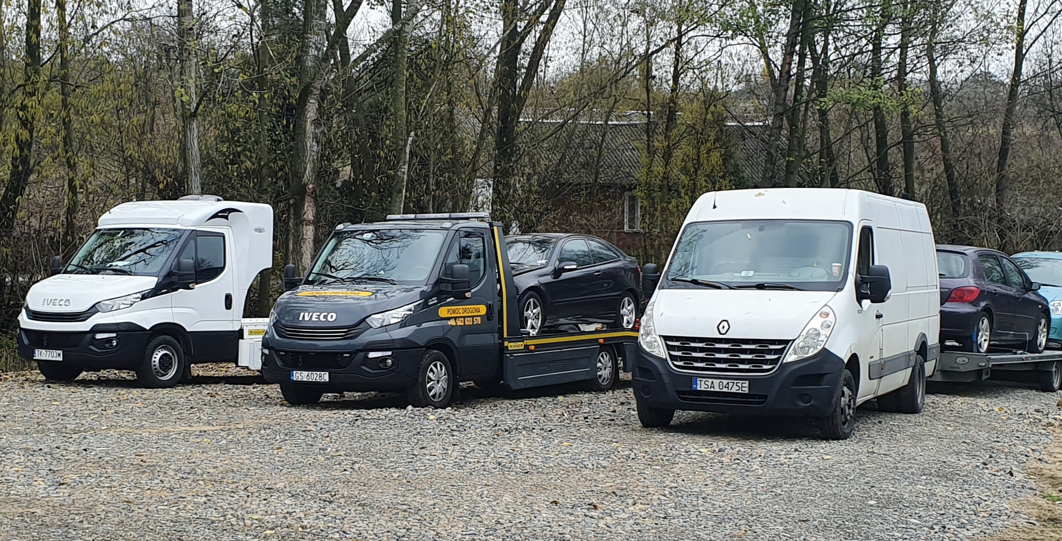 Flota pojazdów pomocy drogowej: biała zabudowa Iveco, czarny Iveco z załadowanym czarnym coupe oraz biały Renault Master z lawetą i dwoma samochodami osobowymi w tle, na szutrowym parkingu.