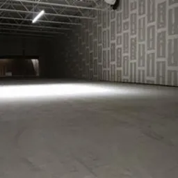 Pusta hala magazynowa z betonową posadzką, surowymi ścianami pokrytymi wzorem i widoczną konstrukcją sufitu z oświetleniem, przygotowana do transportu lub składowania towarów.