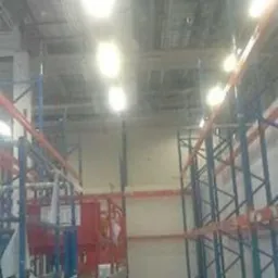 Wnętrze hali magazynowej z zamontowanym oświetleniem sufitowym; widoczne regały magazynowe, podnośnik koszowy oraz instalacje pod sufitem.