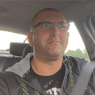 Mężczyzna w okularach, siedzący w samochodzie, ubrany w bluzę z zielonymi sznurkami, patrzy w obiektyw.